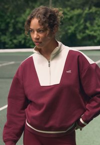 Tumši sarkans un krēmkrāsas džemperis ar rāvējslēdzēju, ģeometrisku krāsu blokveida dizainu, ribveida aprocēm un izšūtu "self." logotipu uz krūtīm.