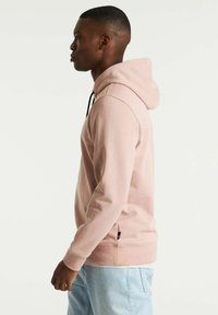Hoodie rosa chiaro con tasca frontale, cordini neri, polsini e orlo a costine. Abbinato a jeans azzurri, che mostrano un profilo laterale.