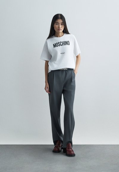 Mladá žena stojí proti jednoduché zdi, oblečená v bílé tričko Moschino, šedých kalhotách a hnědých botách na podpatku, s rukama v kapsách.