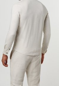 Beige pitkähiha-paita, joka on valmistettu pehmeästä kankaasta. Paidassa on nappihihansuut ja rento leikkaus, yhdistetty samansävyisiin housuihin. Yksinkertainen, tekstuuriltaan hillitty malli.