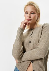 Cardigan beige lavorato a maglia con maniche a coste, scollatura rotonda e bottoni neri. Texture morbida, lunghezza media, con un leggero orlo aperto.