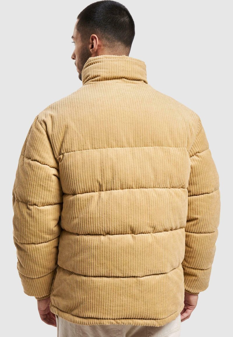 Chaleco de plumas de pana color marrón claro con cuello alto, textura de rayas verticales y secciones acolchadas para aislamiento.