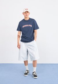 Νέος άνδρας που φοράει μπλε μπλουζάκι Quiksilver, λευκές μακρύ παντελόνες, μαύρα αθλητικά παπούτσια, λευκές κάλτσες και λευκό καπέλο, στέκεται σε μπλε δάπεδο.