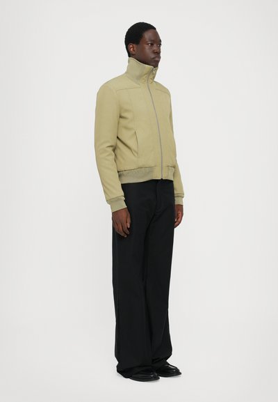 Light olive green zip-up jacket with a high collar, ribbed cuffs, and side pockets, paired with black wide-leg trousers and black shoes.