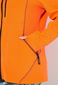 Oranžová zipová bunda s kontrastním vínovým prošíváním a zipy na kapsách. Má dlouhé rukávy a přiléhavý design.