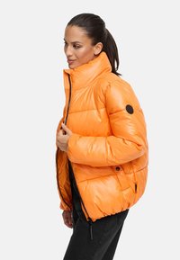 Orange vadderad jacka med hög krage, dragkedjestängning och svart logotypdetalj på ärmen. Slät textur och oversized passform.