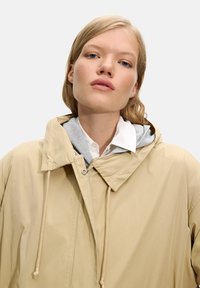 Beige vanntett jakke med stor krage, snor, og grå hettegenser under; glatt tekstur, minimal detaljering, avslappet passform.