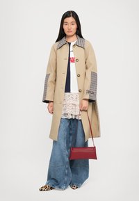 Beige trench coat with a checked collar and patterned cuff accents. Paired with a white lace top, wide denim jeans, and a red handbag.