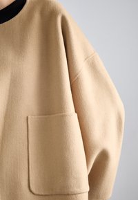 Beige oversize sveter vyrobený z mäkkej látky. Má okrúhly výstrih a jednu prednú vrecko, s viditeľnými švami a voľným strihom.