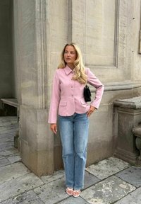 Femme aux longs cheveux blonds portant un blazer rose, un jean bleu, des sandales blanches et tenant un sac à main noir, debout devant un bâtiment en pierre.
