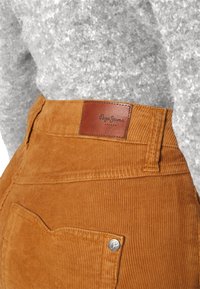 Pantalon en velours côtelé orange brûlé avec poche arrière et écusson logo Pepe Jeans London en cuir marron sur la taille.