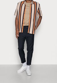 Košile s krátkým rukávem, zapínaná na knoflíky, s vertikálními pruhy v krémové, oranžové a námořnické modři; kombinovaná s námořnickými zúženými kalhotami a bílými slip-on botami.