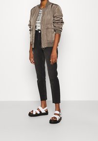 Chaqueta bomber gris con frunces en los hombros, top a rayas, pantalones ajustados negros y sandalias blancas con suela negra y diseño de tiras.