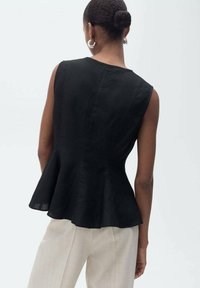 Black peplum top in lightweight fabric, featuring a fitted bodice, short sleeves, and a flared hem. Model wears light beige trousers.