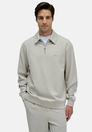 Suéter gris claro con medias cremalleras, cuello plano y un pequeño parche de logo. Presenta mangas largas y un corte relajado.