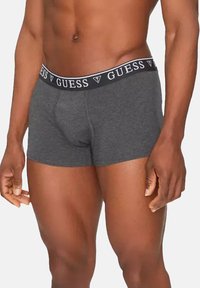 Boxers gris avec un ajustement serré, fabriqués en tissu doux. Dotés d'une taille noire avec le logo "GUESS" en blanc.