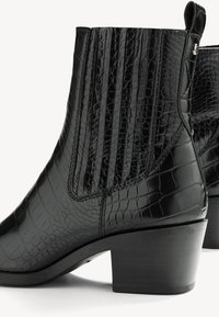 Botines negros con textura de cocodrilo, presentan costuras verticales y un tacón cuadrado. Fabricados en material sintético con un acabado suave.