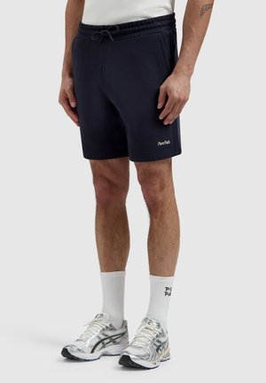 Man draagt marineblauwe Pure Path-shorts, witte hoge sokken met zwarte tekst en zilveren sportschoenen, staand tegen een effen lichte achtergrond.