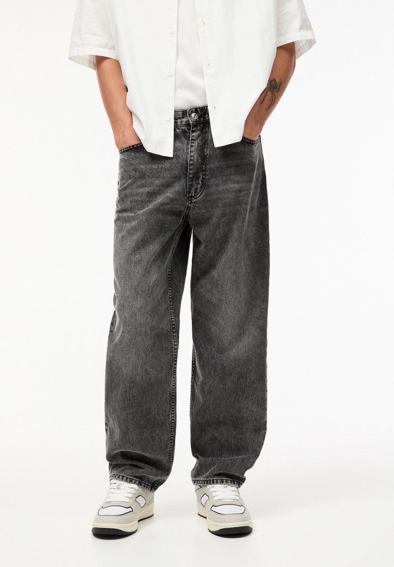 Personne portant un jean noir ample, des baskets blanches, une chemise blanche, et une chemise blanche à manches courtes boutonnée, les mains dans les poches.