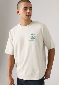T-shirt de manga curta de cor creme, feita de algodão, com um estampado gráfico verde no lado esquerdo do peito com os dizeres "Levi's" e "serving up classics".