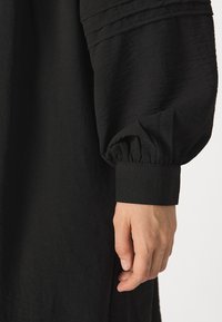 Vestido preto com mangas bufantes, detalhes plissados e punhos ajustados. A textura parece suave e ligeiramente amassada.