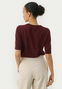 Maglione corto a maniche corte color bordeaux con scollo a giro, abbinato a pantaloni beige chiaro a vita alta, caratterizzati da una texture liscia e da una sartoria curata.