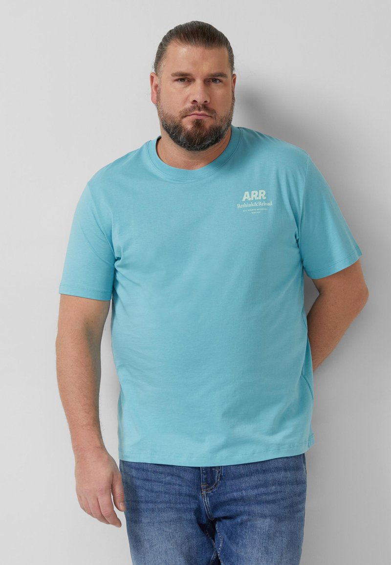 Homme barbu portant un t-shirt bleu clair "ARR Rethink&Reload" et un jean bleu, debout contre un mur gris clair uni.