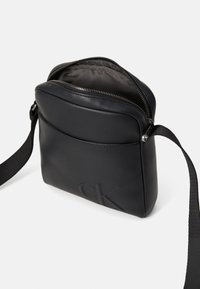 Bolso de cuero negro tipo bandolera con cierre de cremallera, forro interior y logotipo grabado. Presenta una correa plana y una textura suave.