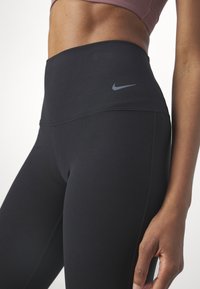 Gros plan d'une personne portant des leggings noirs taille haute Nike et un soutien-gorge de sport mauve, montrant un ventre tonique et la main droite reposant sur le côté.