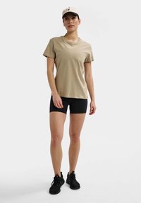 Beige bomulds t-shirt med korte ærmer, rund halsudskæring; parret med sorte shorts og sorte sportssko; person iført en beige kasket.