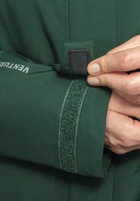 Green outdoor jacket sleeve with a velcro strap; features smooth, matte fabric texture and a subtle branded logo on the sleeve.