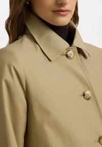 Impermeabile beige con colletto strutturato, caratterizzato da due grandi bottoni di colore chiaro e una texture liscia e opaca.