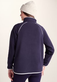 Woman wearing a navy fleece jacket with white piping, dark leggings, and a cream knit beanie, viewed from behind against a plain background.