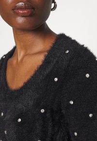 Gros plan d'une personne portant un pull noir pelucheux orné de petits strass dispersés et une boucle d'oreille en argent en forme d'anneau.