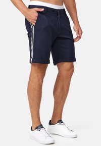Mørkeblå shorts med rullede mansjetter, frontlommer og hvit elastisk midje. Har to vertikale hvite striper langs sidene.
