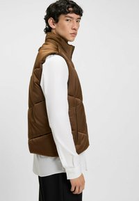 Braune Pufferweste mit hohem Kragen, gestepptem Design und seitlichen Reißverschlüssen. Trägt man über einem weißen Langarmshirt, was den geschichteten Look betont.