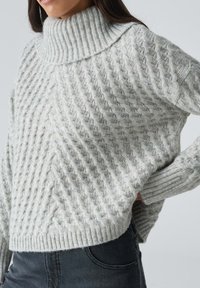 Hellgrauer Strickpullover mit hohem Kragen, der ein strukturiertes Muster und gerippte Bündchen aufweist, kombiniert mit dunkelblauen Jeans.