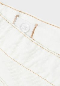 Tessuto bianco con una sottile texture rigata presenta cuciture di un marrone chiaro a contrasto e un bottone trasparente su una piccola linguetta di attacco.