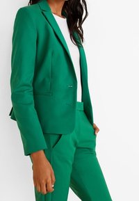 Blazer verde su misura e pantaloni abbinati realizzati in tessuto morbido; presenta un design a un solo petto, rever a punta e tasche laterali.