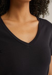 Prim-plan al gâtului și părții superioare a pieptului unei persoane îmbrăcate într-un tricou negru cu decolteu în V, cu părul creț parțial vizibil.