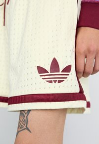 Cremefarvede træningsshorts med perforeret tekstur, udstyret med et maroon i taljen og Adidas-logo i maroon med tre striber design.