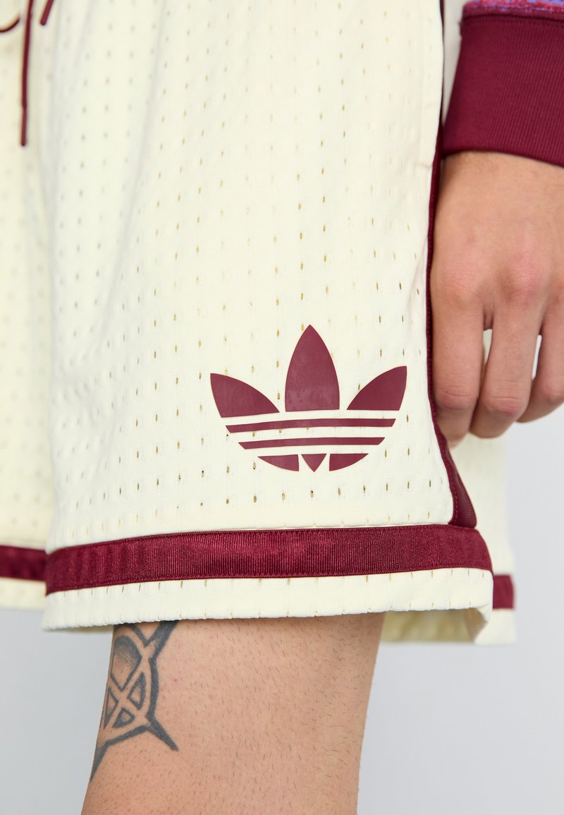 Shorts de sport couleur crème avec texture perforée, dotés d'une taille maroon et d'un logo Adidas en maroon avec un design à trois bandes.