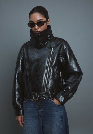 Femme portant une veste en cuir noire surdimensionnée avec un col montant et une ceinture, des lunettes de soleil foncées et un jean bleu, debout devant un fond gris.