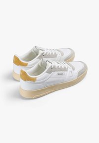 Zapatillas blancas que combinan materiales de cuero y gamuza, con un acabado liso, punta redonda, acentos amarillos en contraste y una suela de goma texturizada.