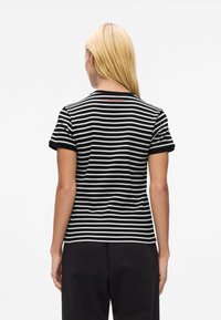 Woman with long blonde hair wearing a black and white horizontal striped short-sleeve shirt and black pants, facing away against a white background.
