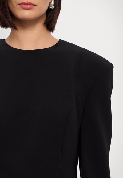 Robe noire avec un col rond et des épaules structurées. Le tissu est lisse avec un corsage ajusté et un design sans coutures. Boucles d'oreilles argentées visibles.