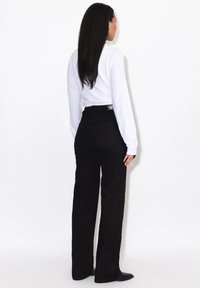 Frau mit langen dunklen Haaren, die ein weißes Langarmshirt und schwarze, hoch taillierte Hosen trägt, steht vor einem schlichten weißen Hintergrund.
