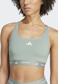Sujetador deportivo verde claro hecho de un tejido suave, que presenta una amplia banda elástica con "TECHFIT" y tres rayas blancas de Adidas en el centro.