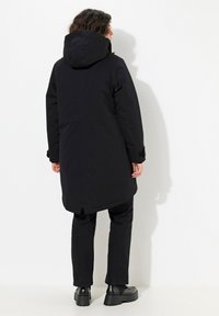 Svart vattentät parkas med huva, långa ärmar, justerbara manschetter och en rundad fåll. Bärs med matchande svarta byxor och kraftiga svarta skor.