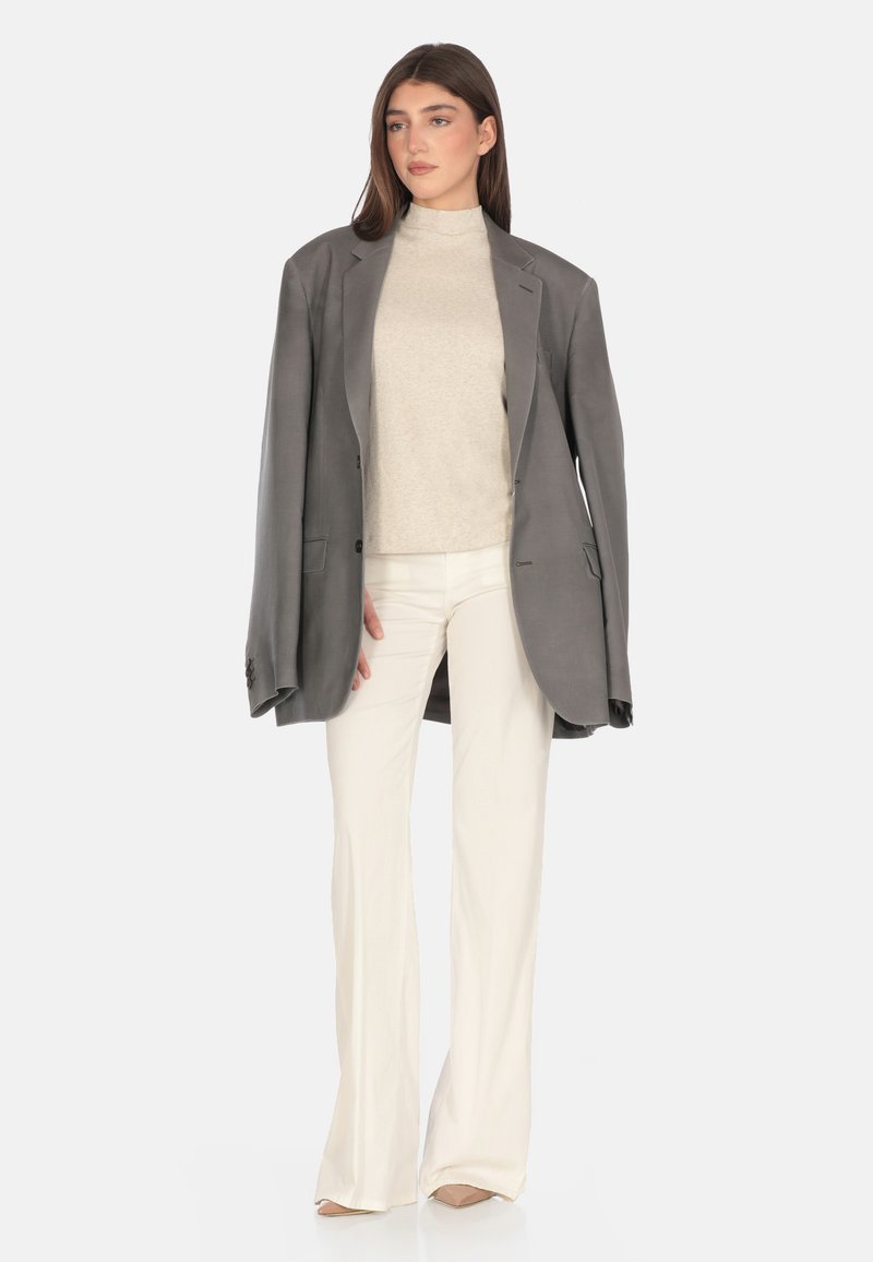 Blazer oversize grigio su un top crema a collo alto, abbinato a pantaloni bianchi a campana e tacchi beige, con tessuti morbidi e design sartoriale.